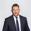 Professional businessman in navy suit and blue tie smiling against neutral background