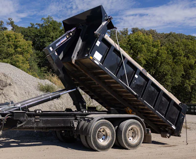 Black dump trailer tilted upright against trees and gravel background