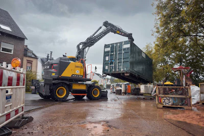 Yellow wheeled excavator moving shipping container at construction site on rainy day