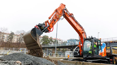 Orange excavator dumping gravel at urban construction site near bridge