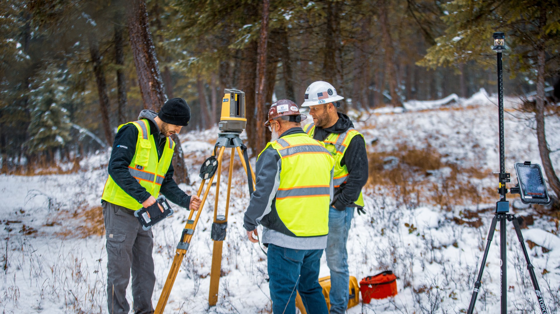 Surveyors in safety vests operating surveying equipment in snowy forest landscape