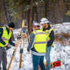 Surveyors in safety vests operating surveying equipment in snowy forest landscape