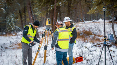 Surveyors in safety vests operating surveying equipment in snowy forest landscape