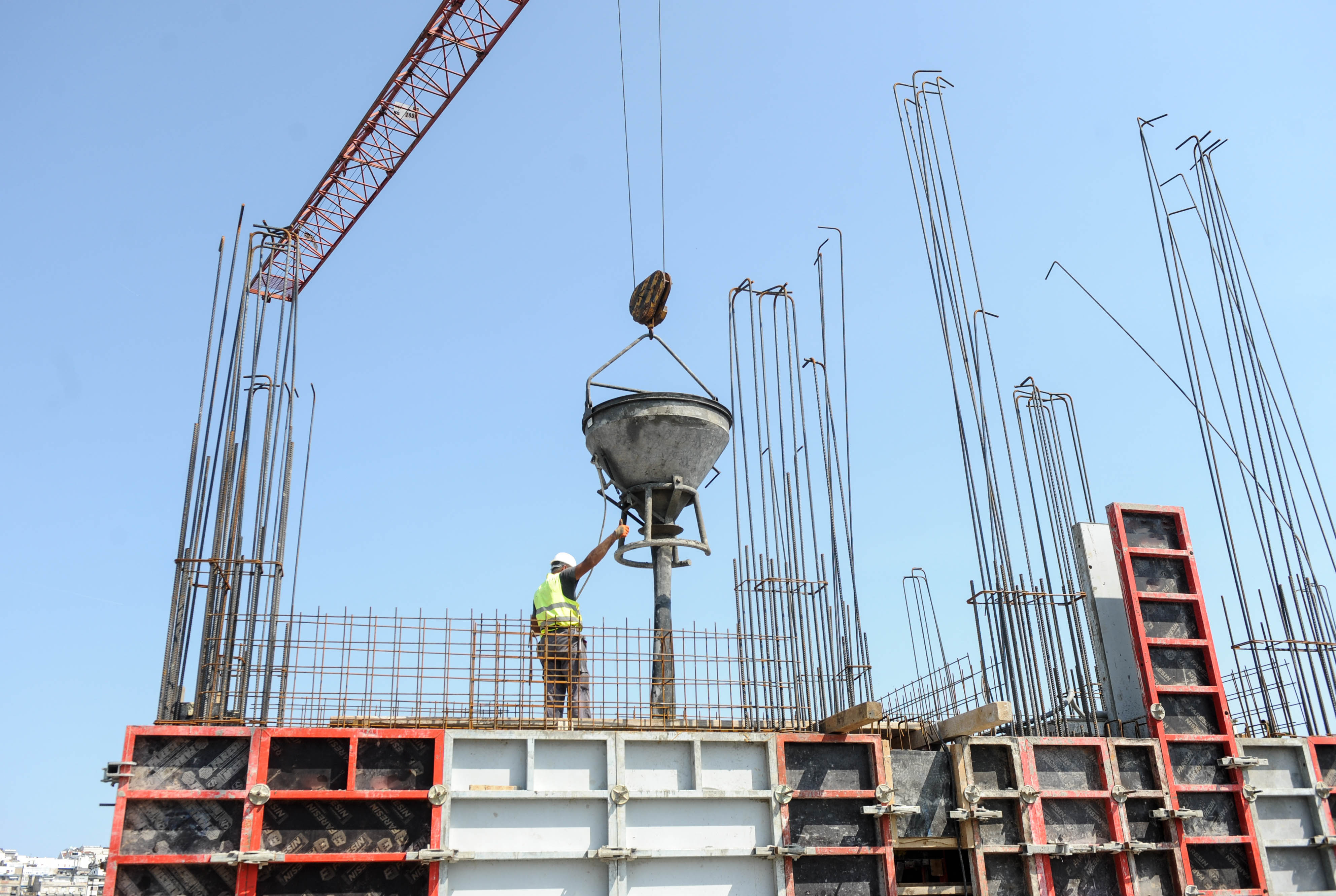 Construction worker guides concrete mixer suspended by red crane at building site with steel reinforcement framework