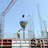 Construction worker guides concrete mixer suspended by red crane at building site with steel reinforcement framework