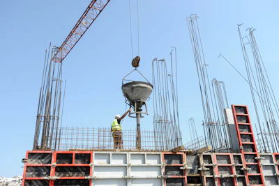 Construction worker guides concrete mixer suspended by red crane at building site with steel reinforcement framework