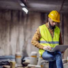 Construction worker in yellow hard hat and safety vest working on laptop at industrial site