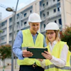 Construction workers in safety vests and hard hats reviewing tablet near apartment buildings