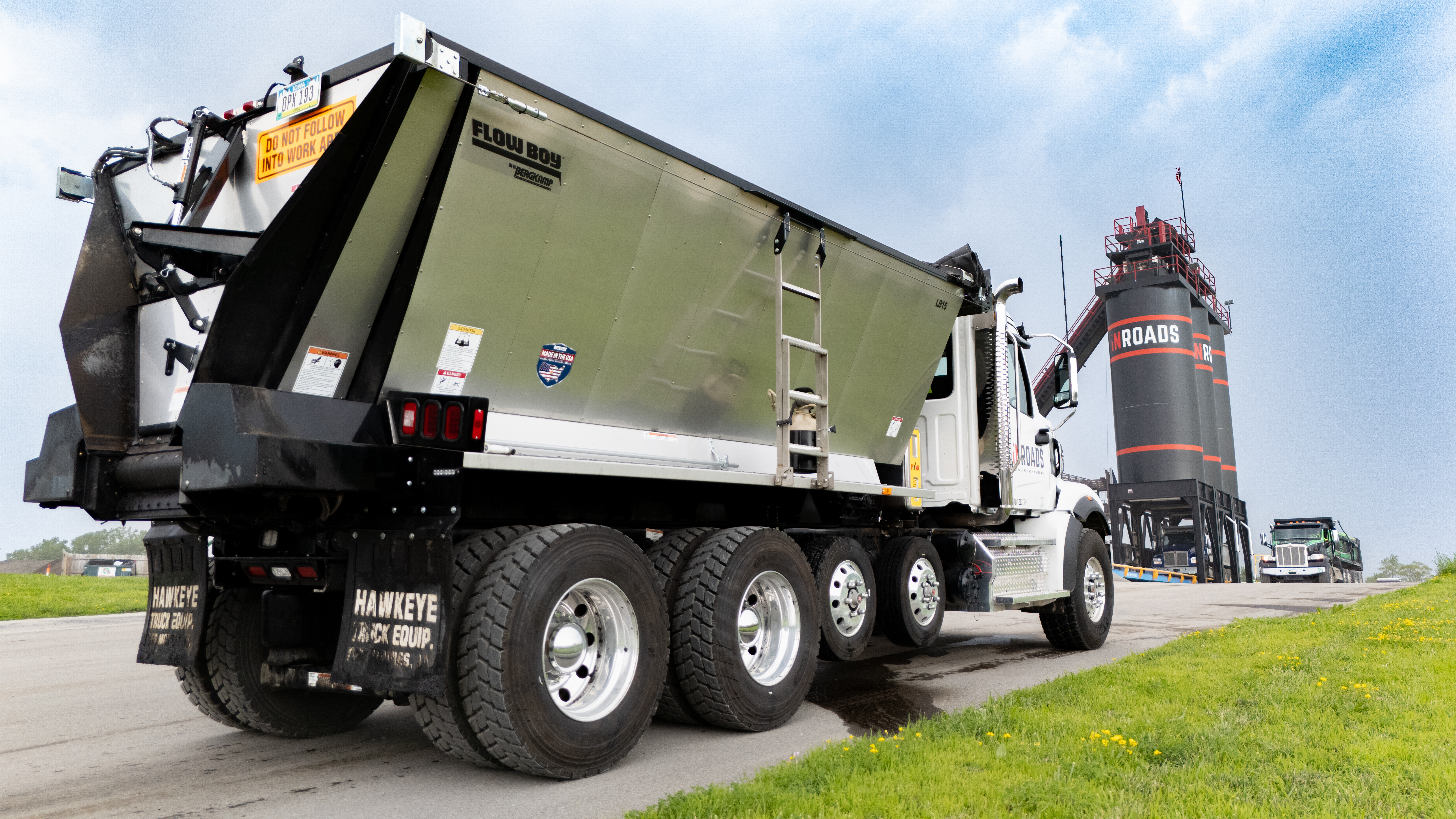 White and olive green commercial dump truck with Hawkeye Gold Group branding on highway with grain silo in background