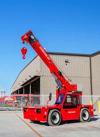 Red Manitou telehandler with extended boom and hook on industrial warehouse lot under clear blue sky