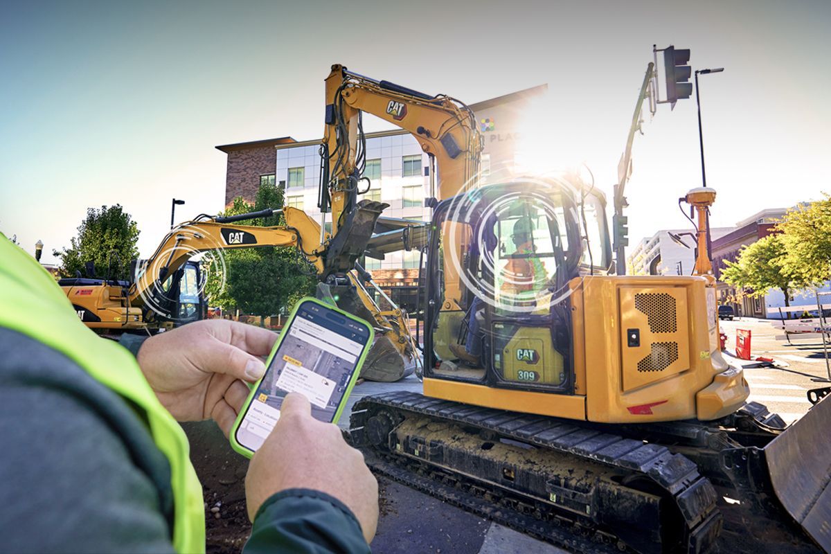 Construction worker holding smartphone monitoring yellow CAT excavator at job site with buildings in background