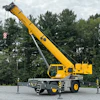 Yellow mobile crane extended with outriggers deployed on gravel lot near pine trees