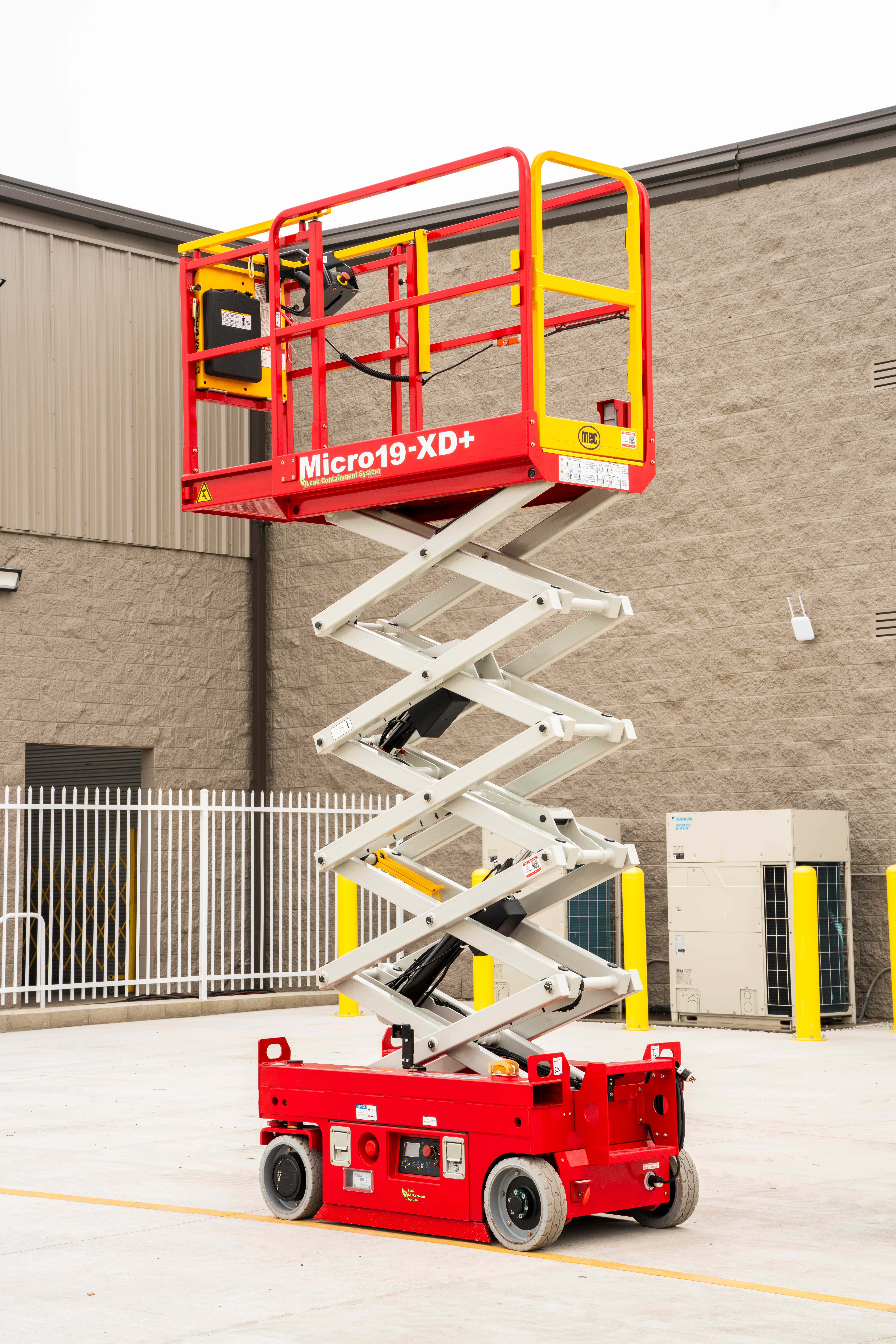Red and yellow Micro19-XD+ scissor lift platform elevated at an industrial facility with concrete building and metal gates
