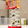 Red and yellow Micro19-XD+ scissor lift platform elevated at an industrial facility with concrete building and metal gates