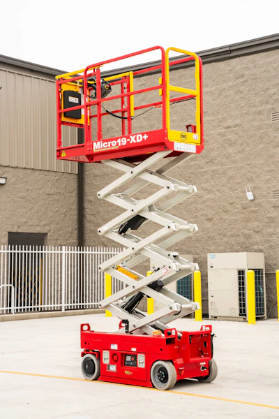 Red and yellow Micro19-XD+ scissor lift platform elevated at an industrial facility with concrete building and metal gates