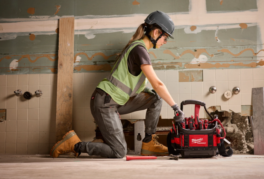 Construction worker in safety vest and helmet kneeling beside red Milwaukee tool bag