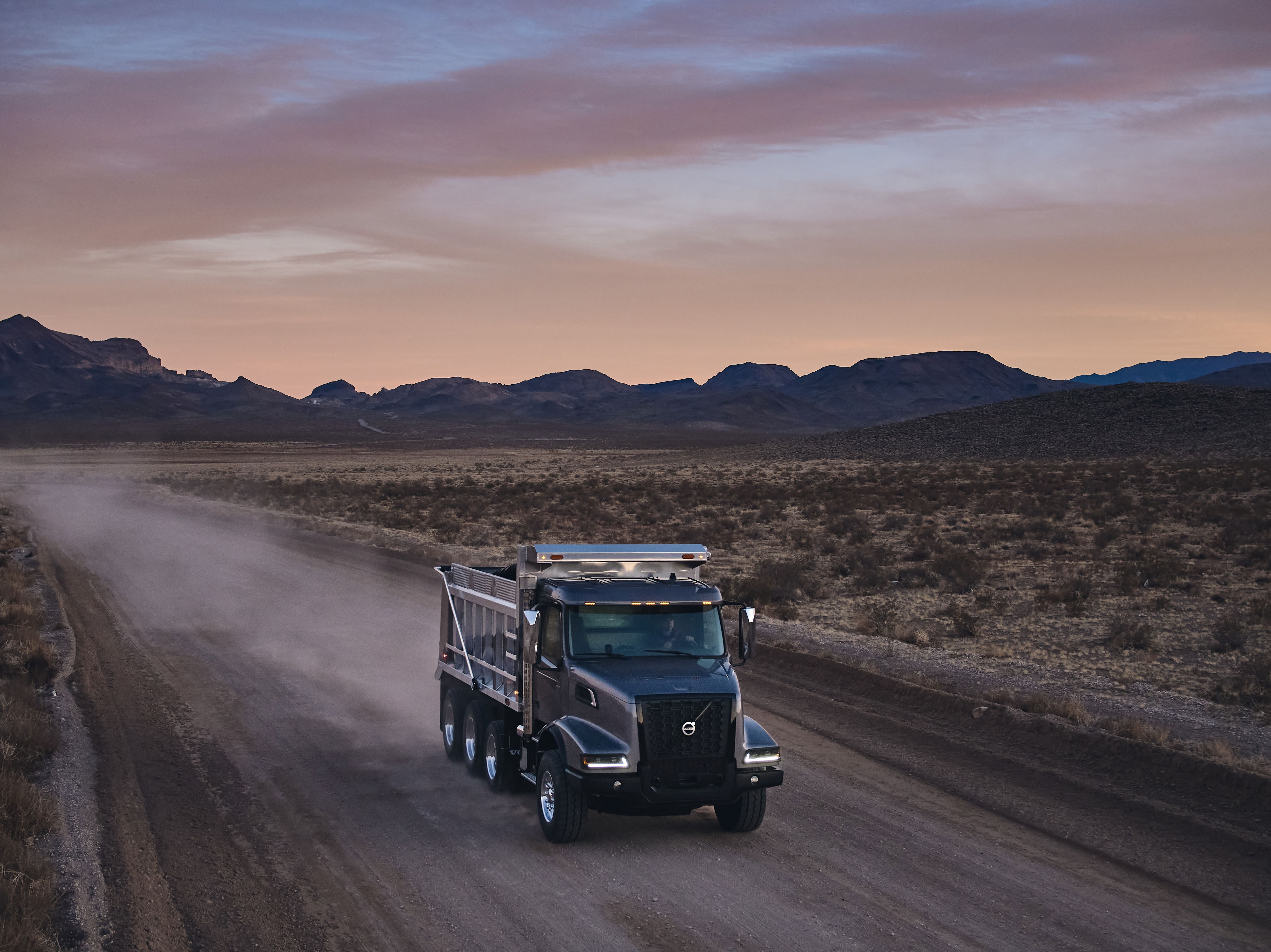 Black Volvo dump truck driving on dusty desert road at sunset with mountains in background
