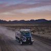 Black Volvo dump truck driving on dusty desert road at sunset with mountains in background