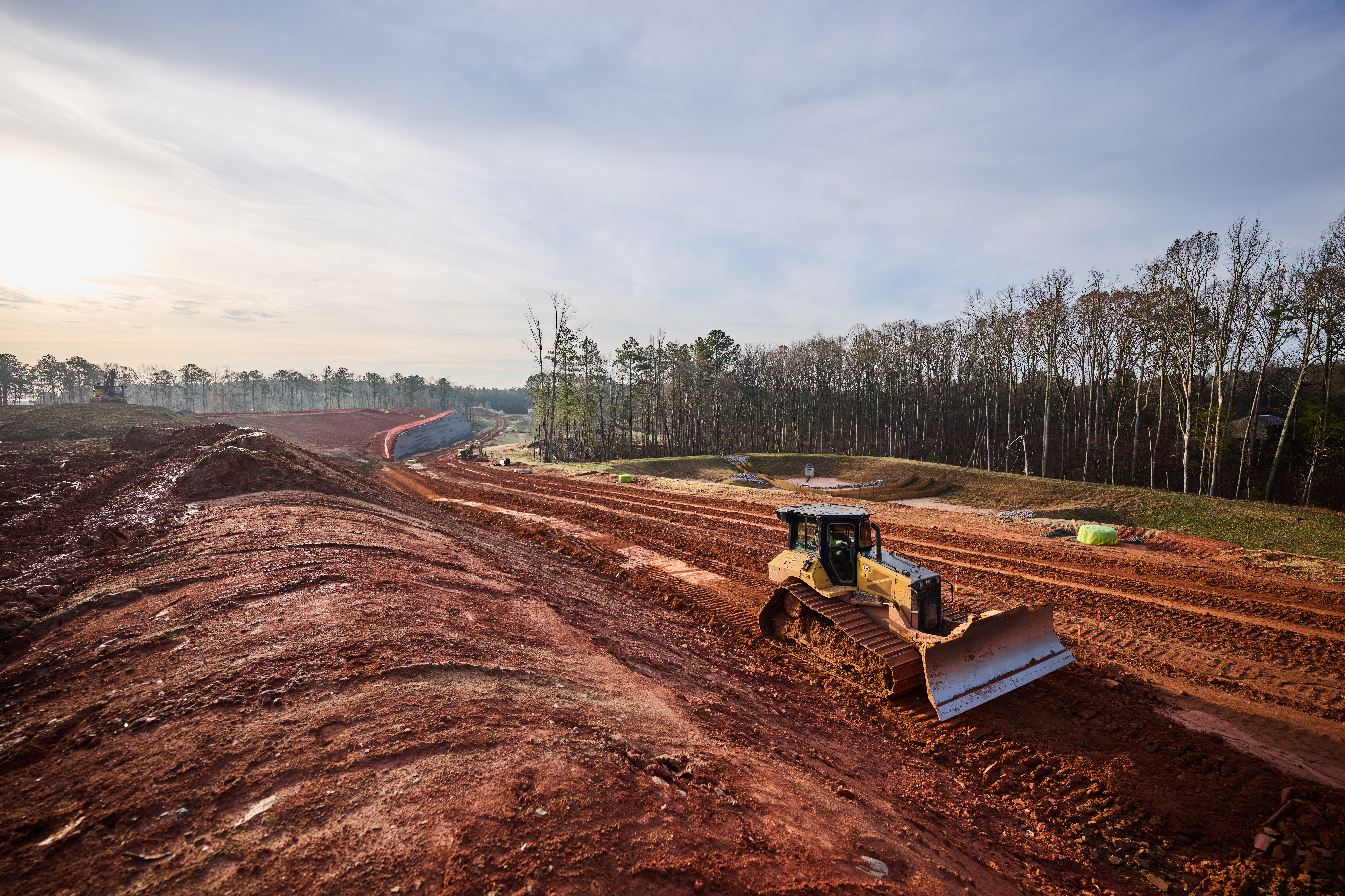 Yellow bulldozer grading red soil on hillside construction site with forested landscape and blue erosion control measures visible