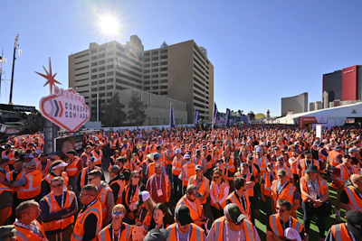 Large crowd of people wearing orange safety vests gathered at outdoor convention with modern buildings and signage in background