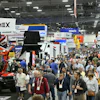 Crowded industrial trade show floor with heavy machinery displays, vendor booths, and attendees wearing identification badges.