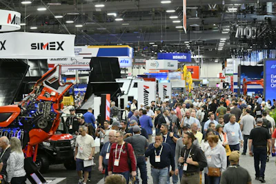 Crowded industrial trade show floor with heavy machinery displays, vendor booths, and attendees wearing identification badges.