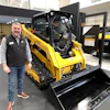 Man standing next to yellow Caterpillar skid steer loader in equipment showroom with additional vehicles displayed