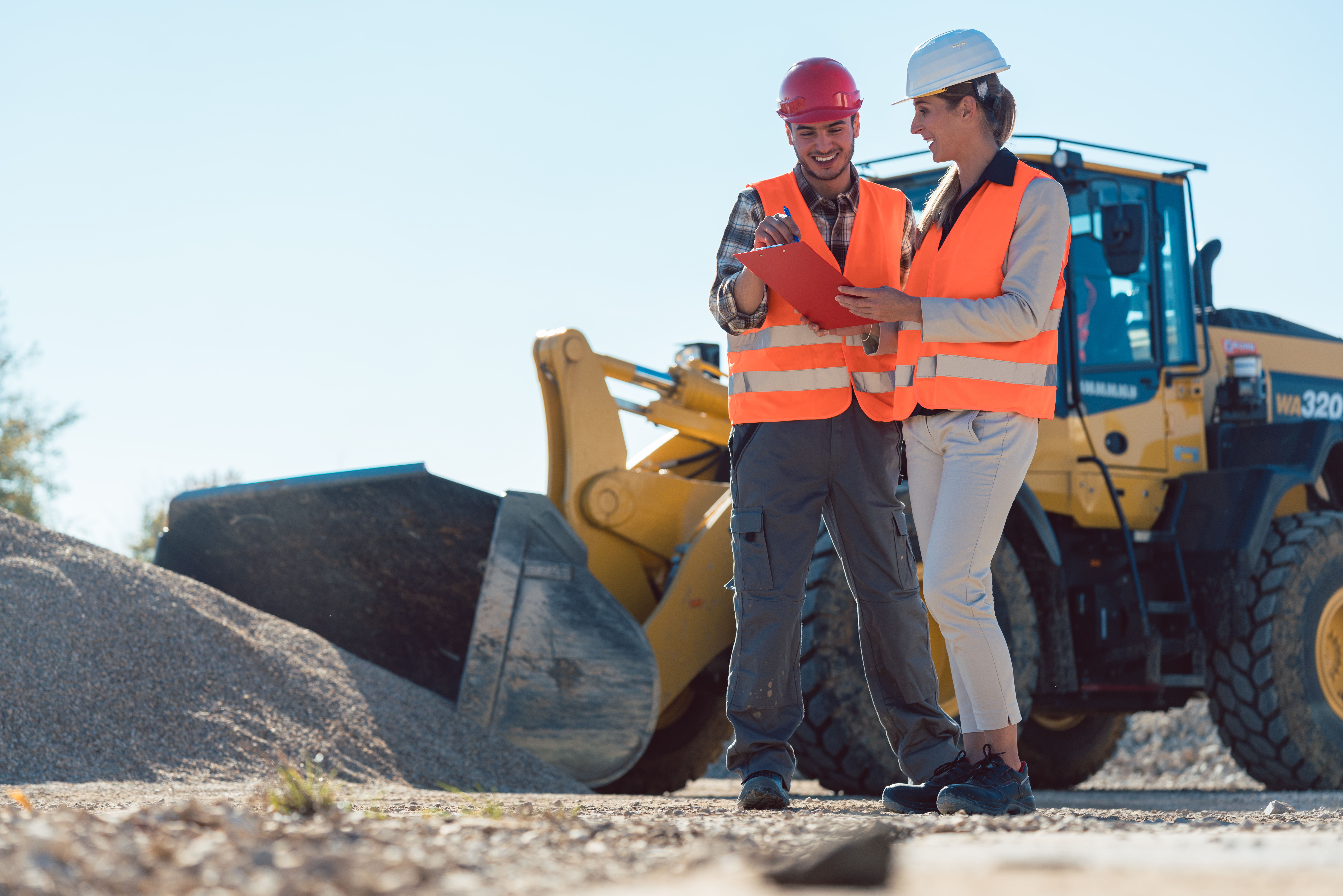 Two construction workers in safety vests review plans at a job site with heavy machinery