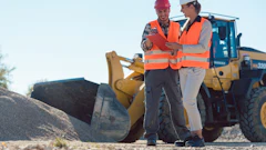 Two construction workers in safety vests review plans at a job site with heavy machinery