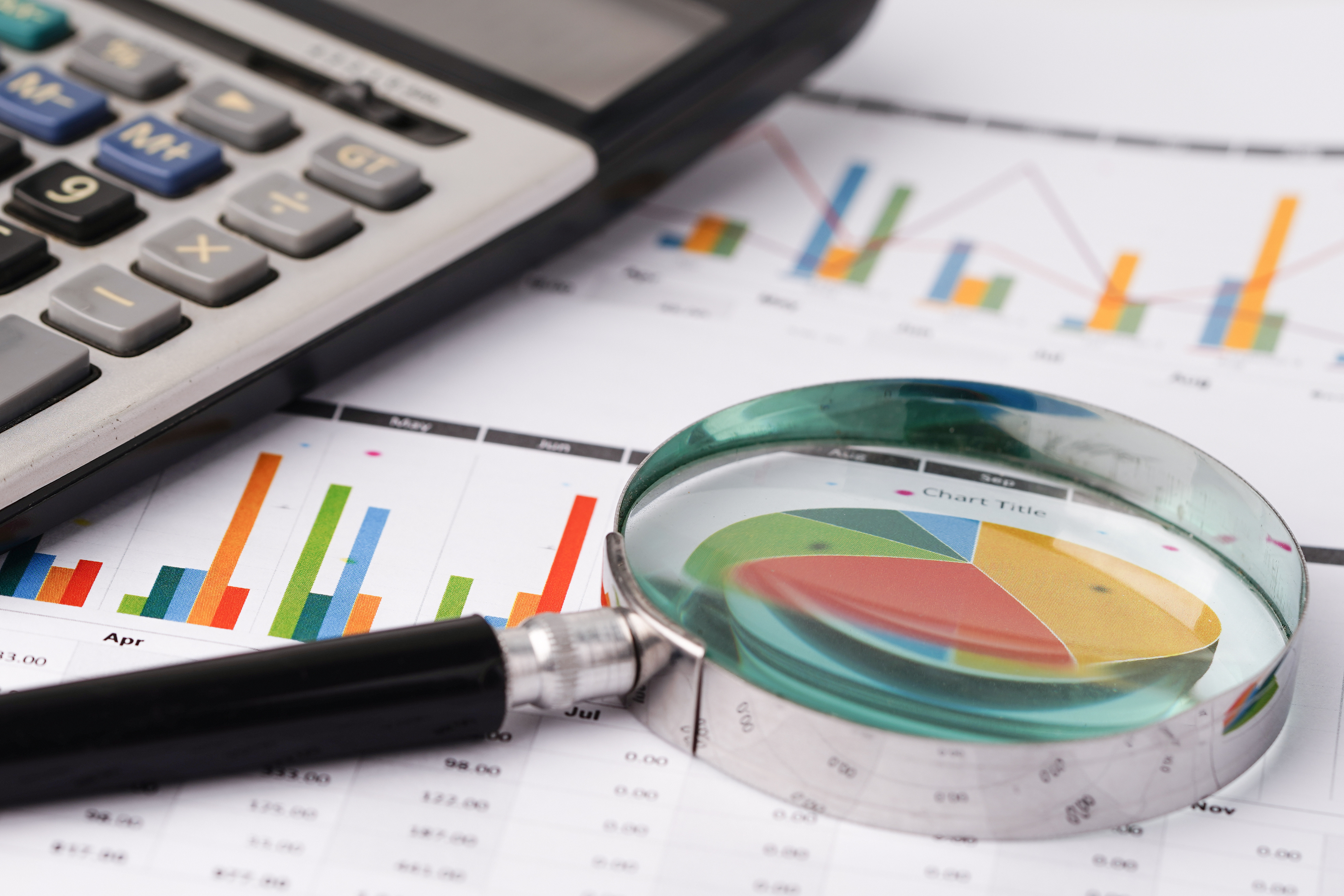 Magnifying glass examining a pie chart surrounded by calculator, bar graphs, and financial data documents