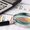Magnifying glass examining a pie chart surrounded by calculator, bar graphs, and financial data documents