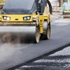 Yellow road roller compactor with headlights compacting asphalt on a construction site with additional equipment in background
