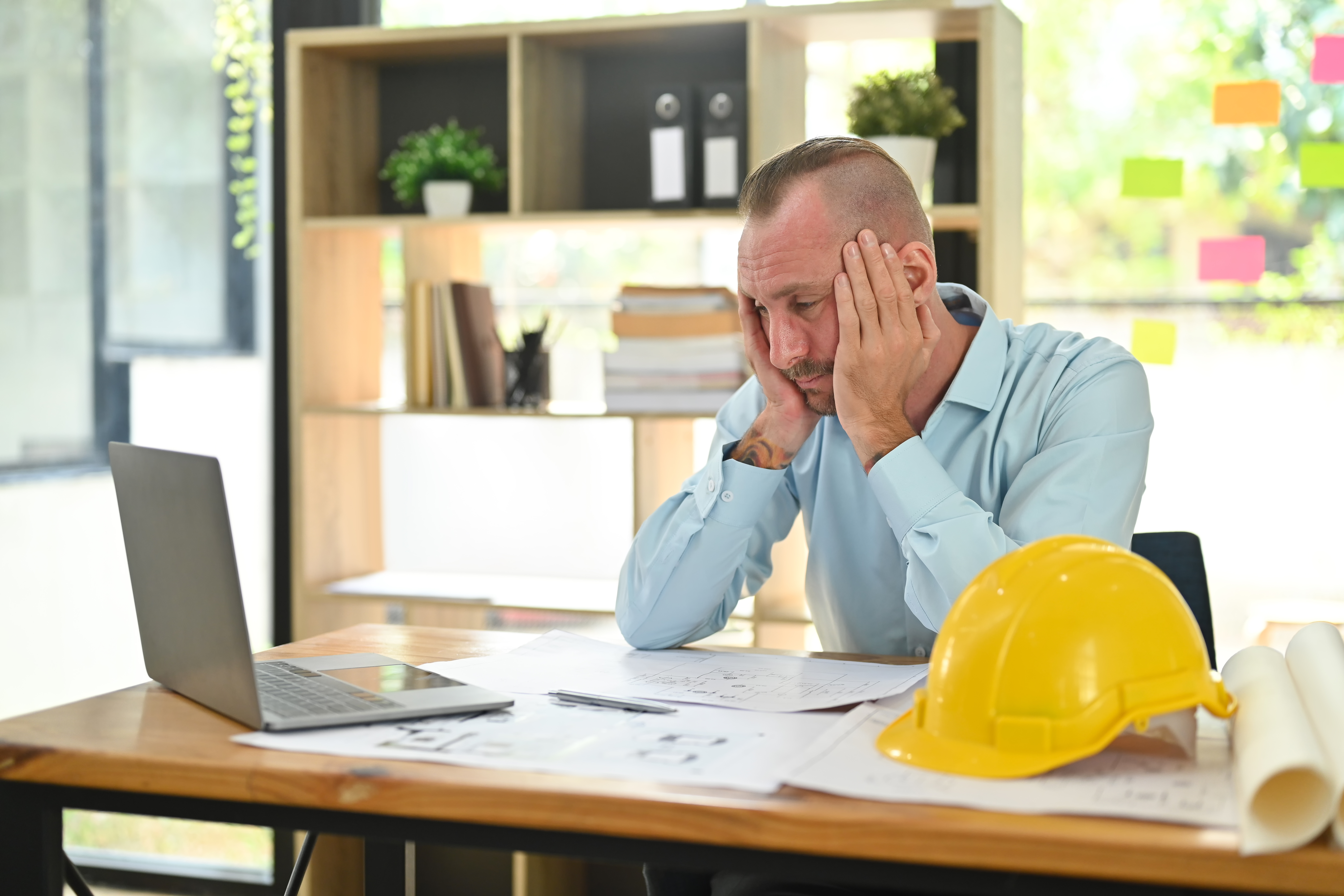 Stressed professional man at office desk with laptop and construction hard hat