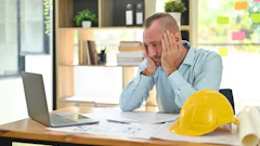 Stressed professional man at office desk with laptop and construction hard hat