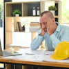 Stressed professional man at office desk with laptop and construction hard hat