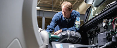 Technician in blue uniform servicing vehicle engine components in an industrial workshop setting