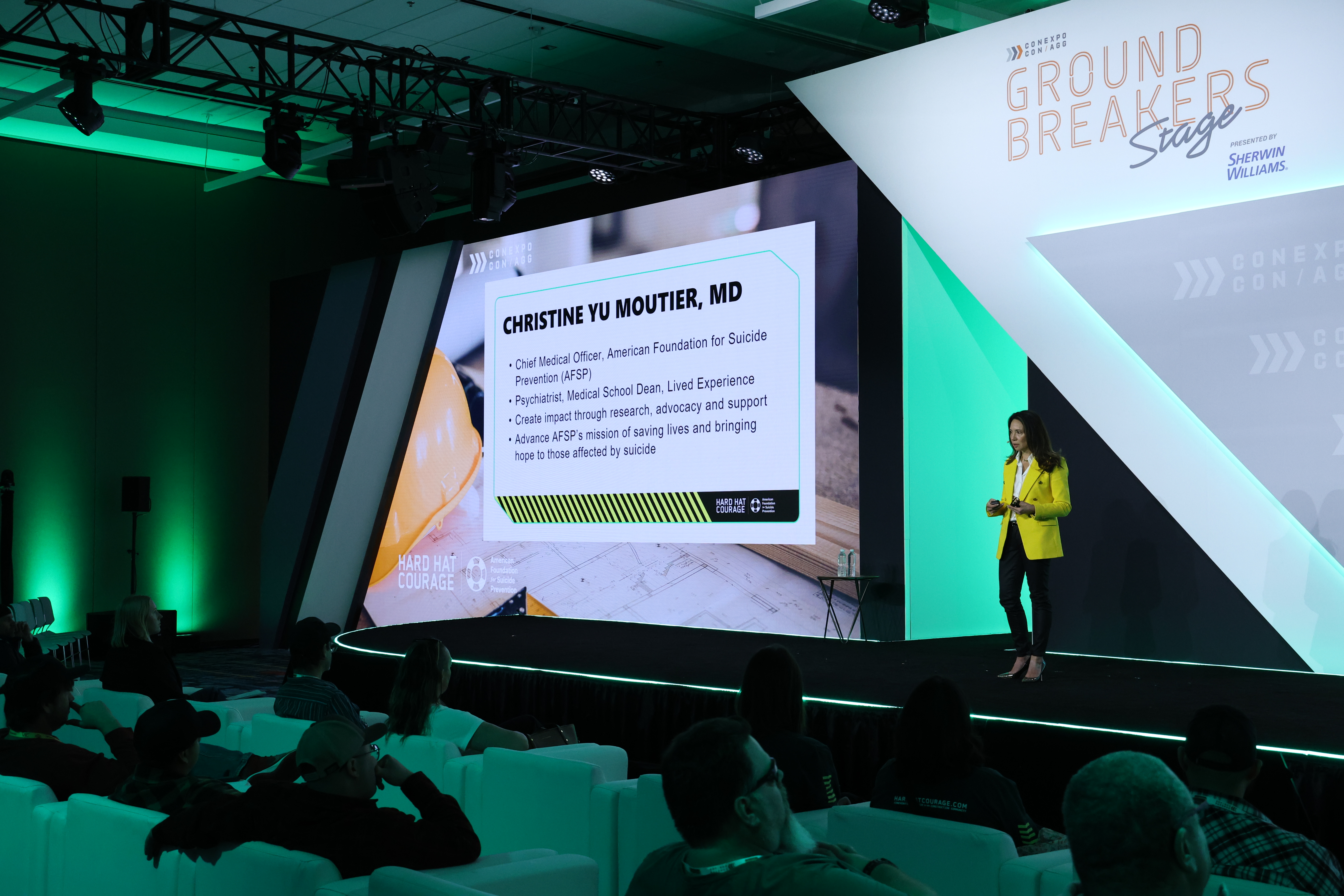 Speaker Christine Yu Moutier, MD presenting at Ground Breakers conference with green-lit stage and projection screen displaying credentials.