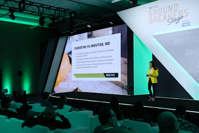Speaker Christine Yu Moutier, MD presenting at Ground Breakers conference with green-lit stage and projection screen displaying credentials.