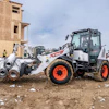 Bobcat telehandler loading metal pipes at construction site with partially built structure and workers