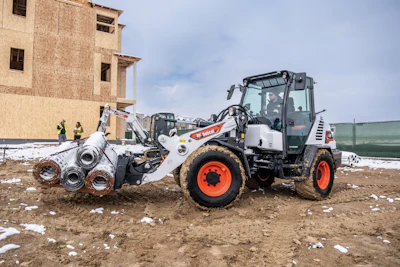 Bobcat telehandler loading metal pipes at construction site with partially built structure and workers