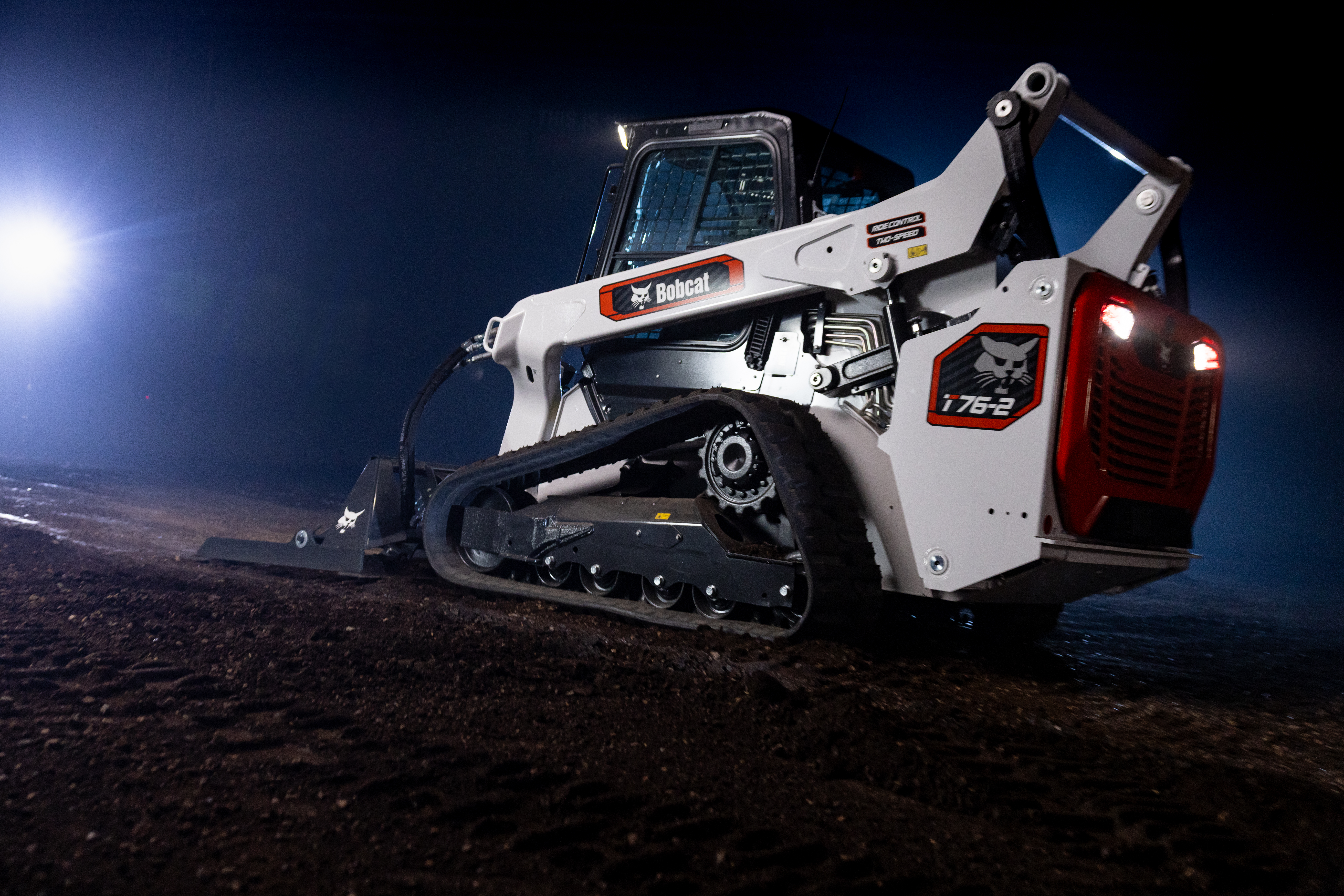 Bobcat T762 compact track loader with white and orange body, illuminated against night sky on dirt terrain
