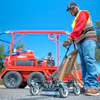 Worker in safety gear operating red industrial spray equipment on sunny day with trees in background