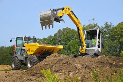Yellow excavator loading soil into dump truck at construction site with green trees in background