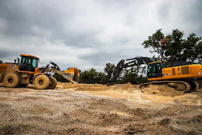 Heavy construction equipment including yellow wheel loader and excavator operating at a dirt job site under cloudy skies