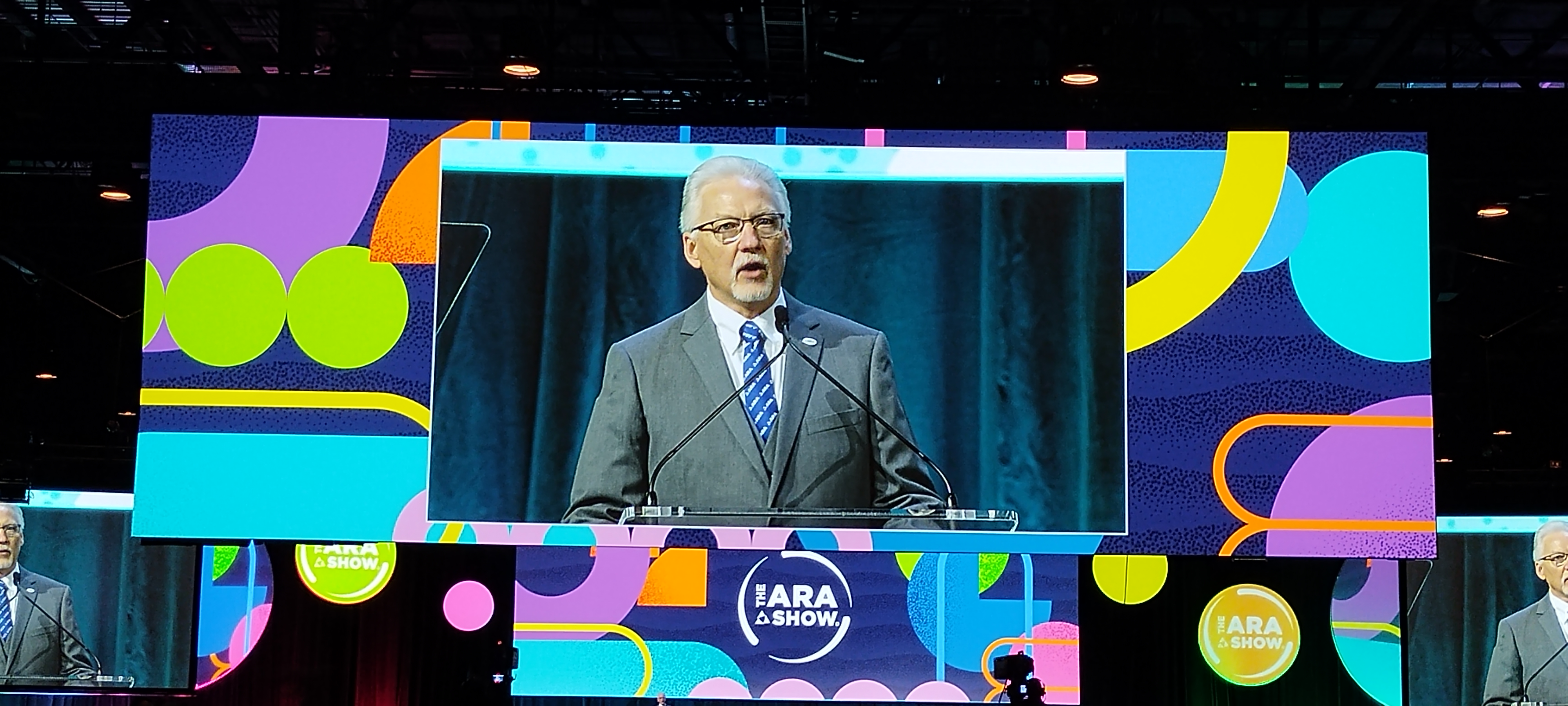 Man speaking at podium during IARA & SHOW conference with colorful geometric branding backdrop