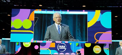 Man speaking at podium during IARA & SHOW conference with colorful geometric branding backdrop