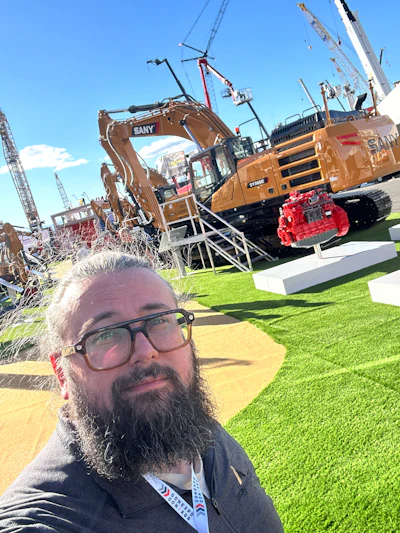 Man with beard and glasses at construction equipment trade show with SANY excavator and machinery in background