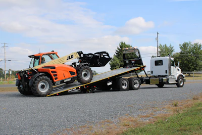 Orange JLG telehandler loaded on white flatbed tow truck on rural highway