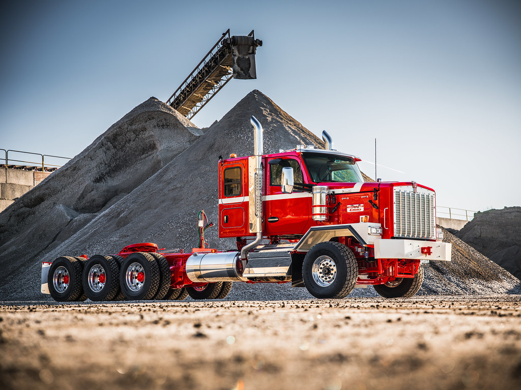 Red and white Peterbilt truck positioned at aggregate quarry with conveyor system and gravel pile in background