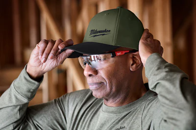 Man wearing green Milwaukee baseball cap and safety glasses in workshop setting